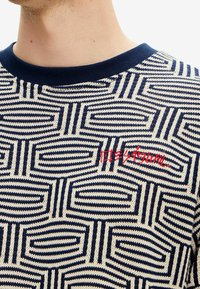 Modrý a krémově vzorovaný pletený svetr s geometrickým designem, zdobený červeným vyšívaným textem. Výrazné jsou kruhové a lineární tvary.