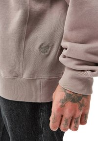 Harmaan vaaleanpunainen huppari, jossa on jalkakuvioidut hihansuut ja helma, ja jossa on brodeerattu logo rinnassa sekä sileä, tekstuuri kangas.