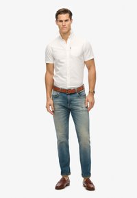 Camicia bianca a maniche corte con taschino, abbinata a jeans leggermente sbiaditi blu chiaro e mocassini in pelle marrone. Cintura marrone inclusa.