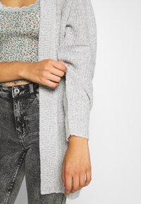 Lichtgrijze gebreide cardigan met een textuurpatroon, open voorkant en lange mouwen, gecombineerd met een bloemmotief top en hoge taille zwarte jeans.