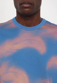 Nahaufnahme einer Person, die ein blau-pink batikgefärbtes T-Shirt mit rundem Ausschnitt vor einem einfarbigen Hintergrund trägt.