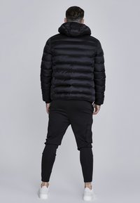 Chaqueta de plumas negra con rayas horizontales, que cuenta con capucha. Combinada con pantalones cargo negros ajustados y zapatillas blancas. Diseño minimalista.