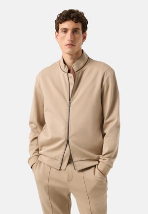 Mann mit lockigem Haar, der eine beige Jacke mit Reißverschluss und passende Hose trägt, mit den Händen in den Taschen, vor einem einfarbigen Hintergrund stehend.