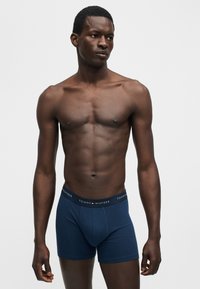 Homme musclé à la peau foncée, debout sur fond blanc, portant un boxer bleu marine Tommy Hilfiger, regardant vers sa droite.