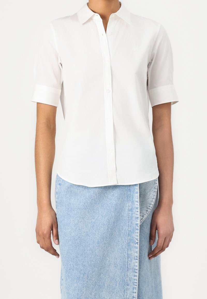 Personne portant une chemise blanche à manches courtes boutonnée et une jupe portefeuille en jean bleu clair, montrée des épaules aux genoux sur un fond uni.