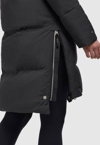 Sort polstret jakke med lynlås i siden delvist åben, hvilket afslører sorte leggings nedenunder, og en hånd afslappet ved siden.