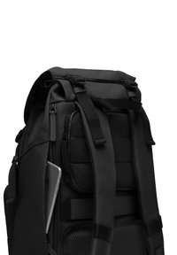 Sac à dos noir avec un design à rabat, comportant des panneaux en maille, des bretelles réglables et une poche latérale contenant un smartphone argenté.