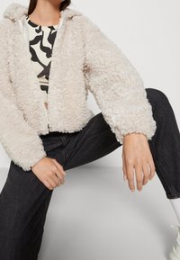 Femme portant une veste à fermeture éclair en sherpa crème, un haut à motifs noir et blanc, un jean noir, des chaussettes blanches, posant jambes écartées sur un fond uni.