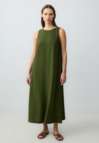 Zielona lniana sukienka bez rękawów o prostym kroju, sięgająca do połowy łydki, zestawiona z brązowymi płaskimi sandałami. Minimalistyczny design i faktura.