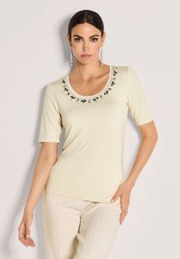 Vrouw met donker haar in een crèmekleurig kortmouwtop met juweelversieringen rond de halslijn en crèmekleurige broek, poserend tegen een effen achtergrond.