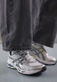 White athletic shoes with silver accents, mesh material, and green detailing. Worn with gray wide-leg denim pants. Ground view.