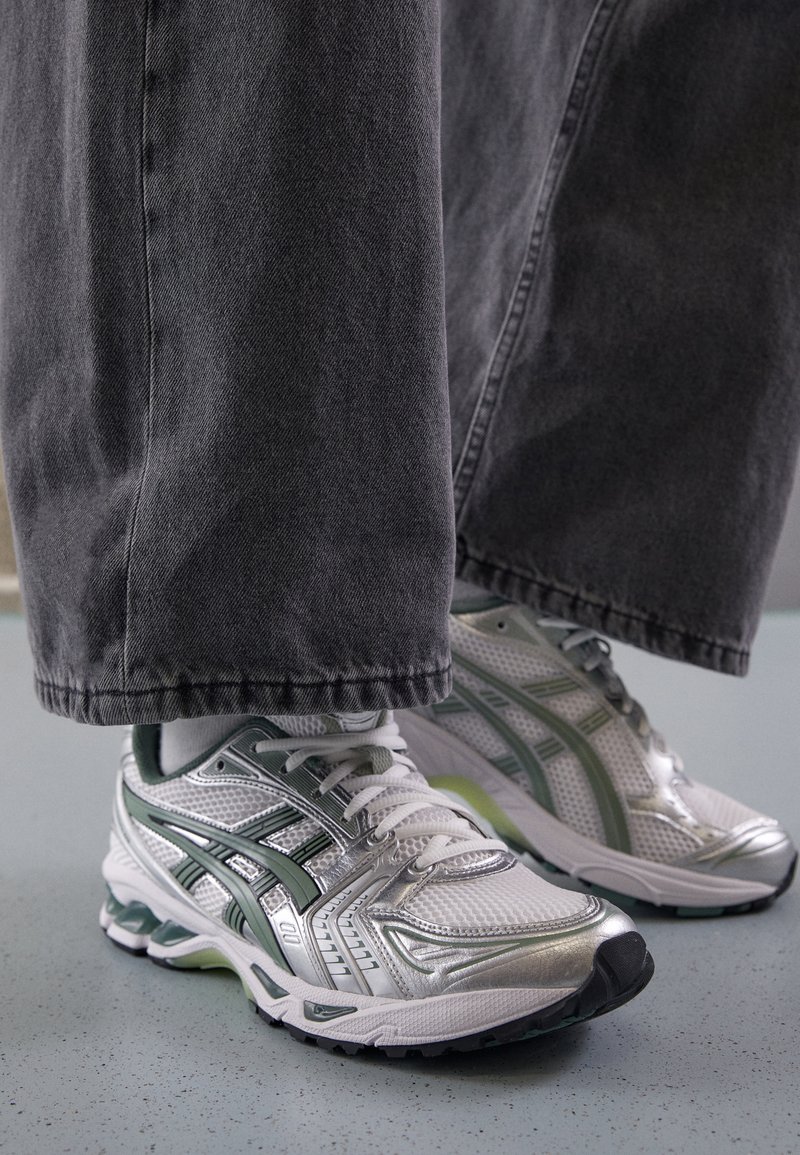 White athletic shoes with silver accents, mesh material, and green detailing. Worn with gray wide-leg denim pants. Ground view.