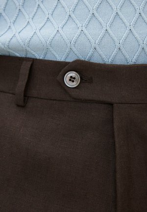 Vaaleansininen, teksturoitu neulepaita, joka on työnnetty tummanruskeiden housujen sisään. Housut on kiinnitetty yhdellä nelireikäisellä napilla.