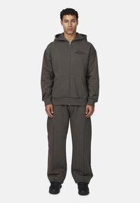 Felpa grigia con zip, tasche frontali e cappuccio, abbinata a pantaloni grigi coordinati con dettagli di cucitura laterale e sneakers nere.