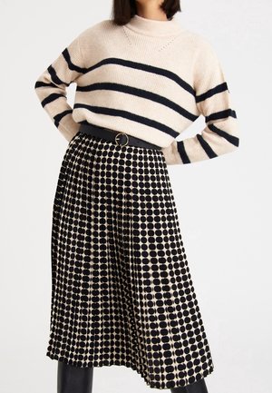 Frau trägt einen cremefarbenen Pullover mit schwarzen horizontalen Streifen, der in einen schwarzen und beigen, gepunkteten Faltenrock mit schwarzem Gürtel gesteckt ist.