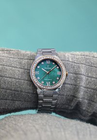 Stříbrné nerezové hodinky se zeleným ciferníkem, římskými číslicemi, diamantovými akcenty a texturovaným páskem, odpočívající na šedém textilním pozadí.