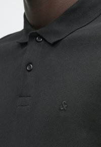 Gros plan sur une personne portant un polo noir à col avec deux boutons et un petit logo brodé «&f» sur la poitrine.