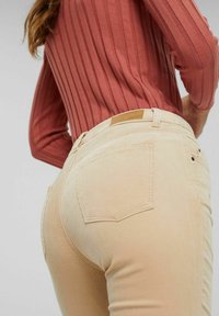 Femme portant un pantalon en velours côtelé beige et un haut à manches longues côtelé couleur rouille, vue de dos et de la taille vers le bas.