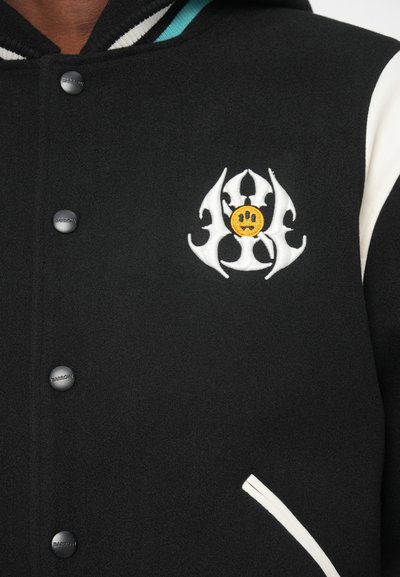 Schwarze Collegejacke mit weißen Akzenten, die auf der Brust ein grafisches Motiv eines lächelnden Gesichts umgeben von abstrakten Formen zeigt. Strukturierter Stoff und Druckknöpfe.