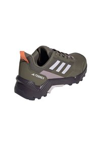 But sportowy Terrex w oliwkowej zieleni z białymi paskami. Posiada chropowatą, materiałową cholewkę, pomarańczową zakładkę na pięcie oraz wytrzymałą czarną podeszwę.
