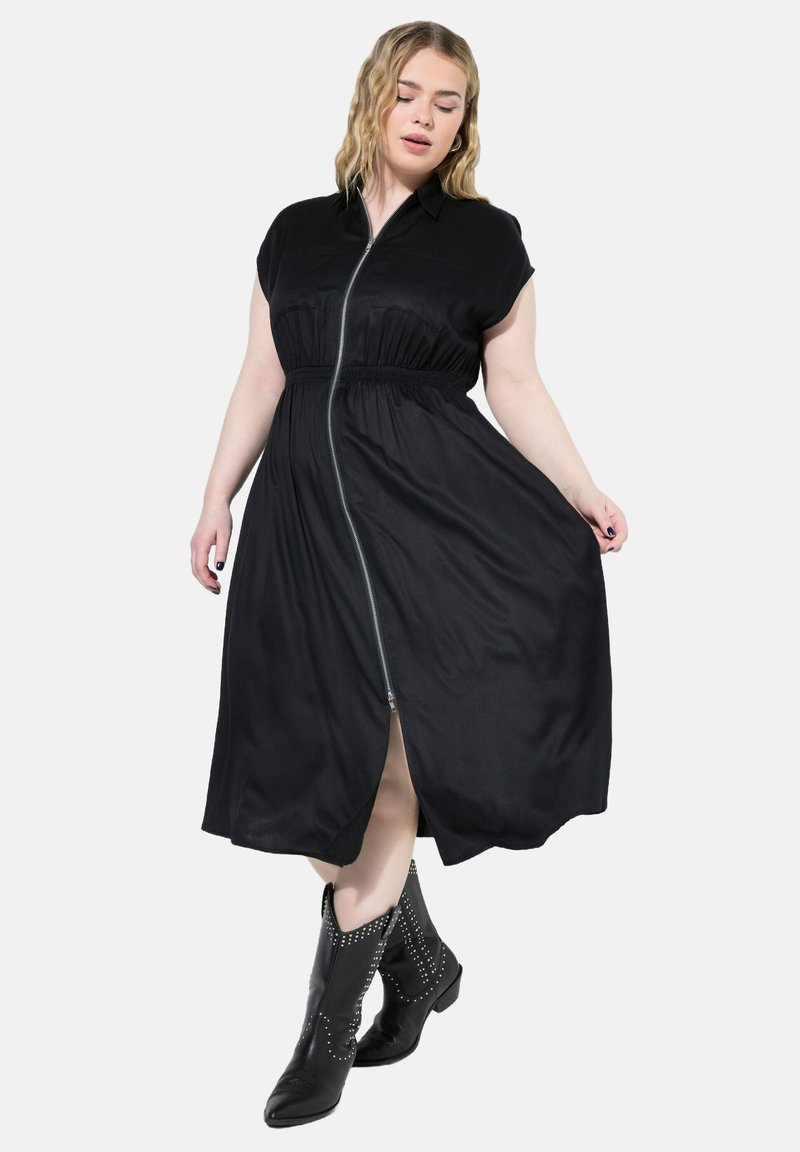 Vestito midi nero con maniche corte, chiusura frontale con zip, vita elastica e spacchi laterali. Indossato con stivali da cowboy neri con borchie.