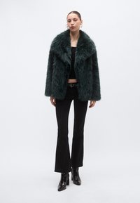 Abrigo de piel sintética verde con cuello ancho, combinado con un top corto negro y pantalones acampanados. La modelo lleva botas de tacón alto. Se aprecia una textura suave.