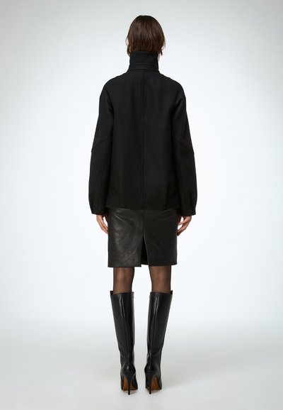 Sort uldfrakke med høj krave, oversized ærmer og en figursyet silhuet, kombineret med en sort læder pencilnederdel og knæhøje støvler.