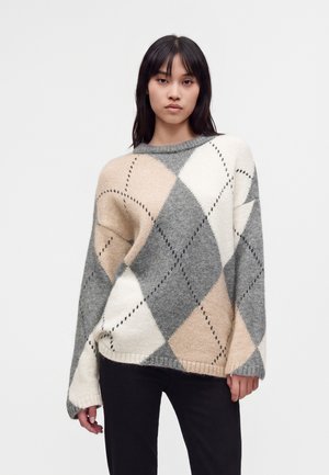 Jeune femme portant un pull ample à motif losanges beige, gris et blanc avec des lignes noires en pointillés, associé à un pantalon noir.