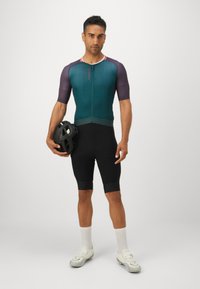 Tenue de cyclisme pour hommes comprenant un haut à manches courtes de couleur bleu sarcelle foncé et violet, associé à un short noir, fabriqués en tissu lisse et extensible. Tenant un casque noir.