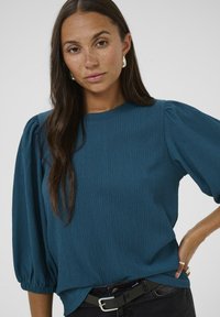 Haut texturé en vert d'eau avec des manches bouffantes et un col rond, associé à un jean noir et une ceinture noire, avec une finition brillante.