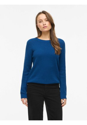 Donna che indossa un maglione lavorato a maglia blu testurizzato e pantaloni neri, in piedi davanti a uno sfondo bianco uniforme con un'espressione neutra.
