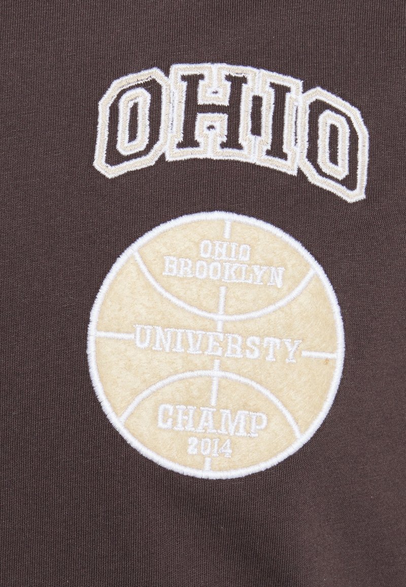 Tissu marron avec un texte brodé "OHIO" au-dessus d'un ballon de basket beige avec l'inscription "OHIO BROOKLYN UNIVERSITY CHAMP 2014" cousue à l'intérieur.
