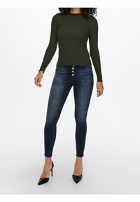 Jean skinny bleu foncé avec fermeture à cinq boutons à l'avant, associé à un haut côtelé vert olive ajusté à manches longues. Textures lisses, coupe sur mesure.