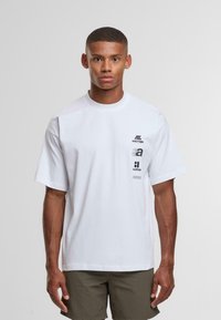 Joven con el cabello corto vistiendo una camiseta blanca de gran tamaño con diseños de texto en negro y pantalones cortos verde oliva, de pie contra un fondo sencillo.