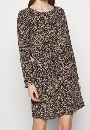 Kvinde iført en langærmet knælængde kjole med et sort og beige abstrakt mønster og en indsnøret talje.