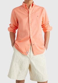 Mann trägt ein langärmliges, korallenfarbene Hemd mit hochgekrempelten Ärmeln und cremefarbene, knielange Shorts, Hände in den Taschen.