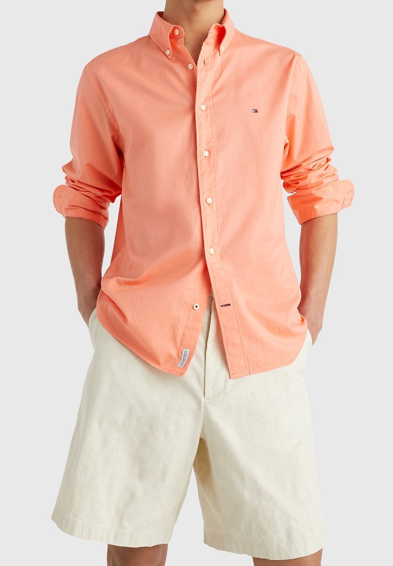 Mann trägt ein langärmliges, korallenfarbene Hemd mit hochgekrempelten Ärmeln und cremefarbene, knielange Shorts, Hände in den Taschen.