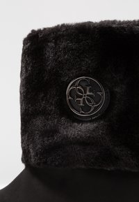 Bota negra con puño de pelaje suave y acolchado, y un emblema metálico redondo con letras entrelazadas en el lado externo.