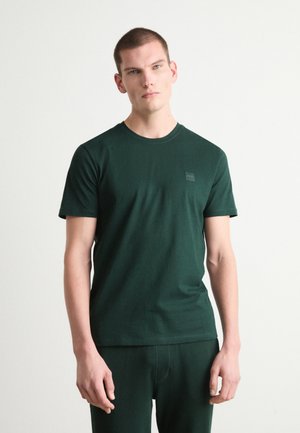 Camiseta de algodón verde con cuello redondo y mangas cortas, que presenta un pequeño parche con logo en el pecho. Textura suave y diseño ajustado.