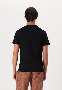 Black short-sleeve t-shirt with a crew neck, viewed from the back, paired with brown patterned pants featuring a drawstring waist.