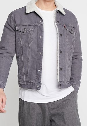 Homme portant une veste en jean grise avec col en polaire blanche sur un t-shirt blanc et un pantalon gris, debout avec une main dans la poche.