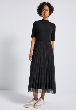 Woman with curly hair wearing a black short-sleeve top and patterned pleated black midi skirt paired with white sneakers against a plain background.