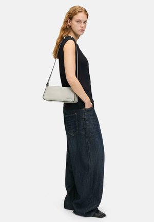 Kleine graue Lederhandtasche mit langem Tragegurt, runder Form, glatter Textur, kombiniert mit einem enganliegenden schwarzen Oberteil und weit geschnittenen dunklen Jeans.