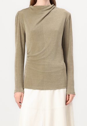 Vrouw draagt een beige lange-mouwen fluwelen top met subtiele plooien aan de linkerkant, gecombineerd met een witte rok.