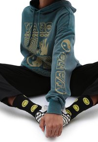 Felpa color teal con grafiche gialle, maniche lunghe e tasca frontale. Pantaloni neri e calze a fantasia con faccine sorridenti gialle e logo Vans.