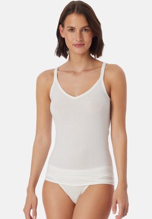 Frau mit schulterlangem braunem Haar, die ein weißes ärmelloses Camisole und passende Unterwäsche trägt, vor einem schlichten Hintergrund stehend.