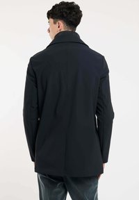 Zwarte jas met een gestructureerde kraag, gladde stof en getailleerde pasvorm. Beschikt over een rugventilatie en subtiele stikseldetails. Draagt over donkere broeken.