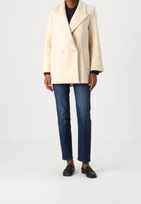 Dubbelzijdig crèmekleurige wollen jas met inkepingen, voorzien van zijzakken en een losse pasvorm, gecombineerd met donkerblauwe jeans en zwarte loafers.