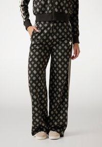 Sorte mønstrede træningsbukser med hvide geometriske designs, brede ben og sidelister, parret med lyse slip-on sko.