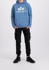 Felpa con cappuccio blu con logo bianco "Alpha Industries", abbinata a pantaloni cargo neri con più tasche e sneakers nere.